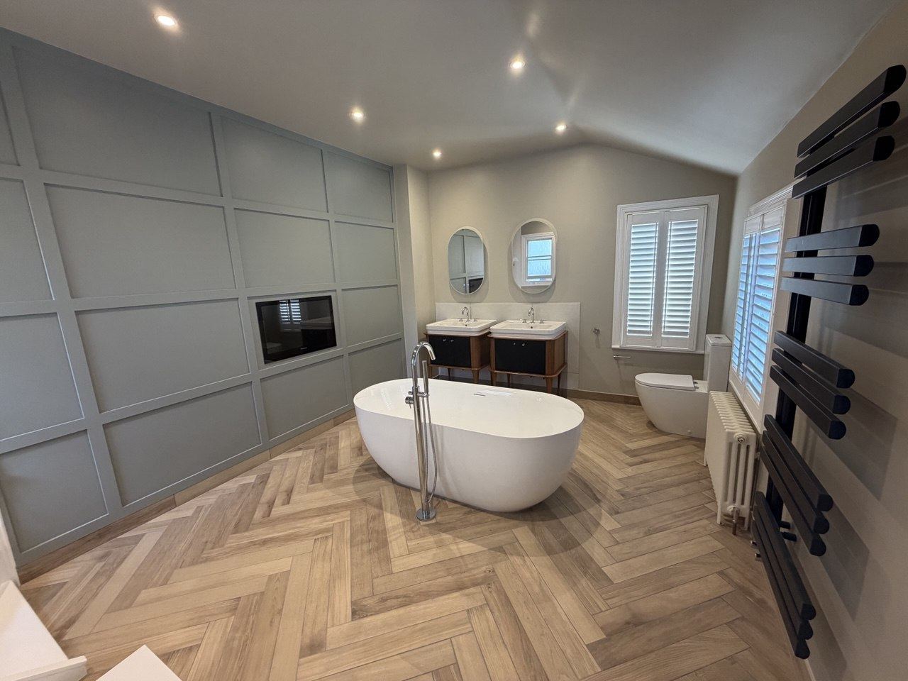 A bathroom with a freestanding bath, twin washstands with oval mirrors and a herringbone tile floor