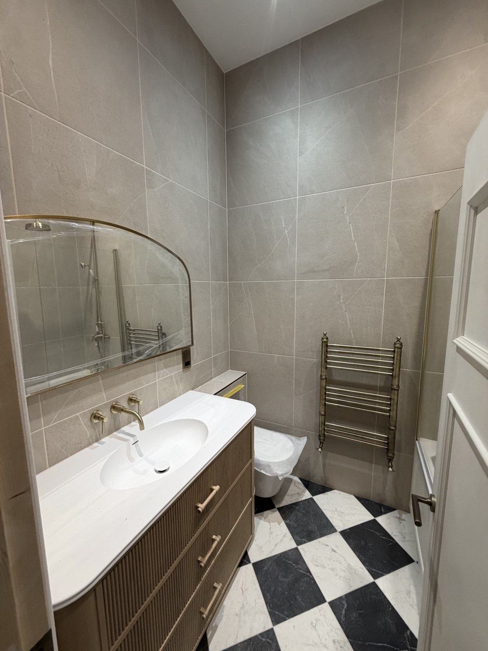 A bathroom with a black-and-white chequerboard floor, a fluted vanity, brass taps and large-format wall tiles