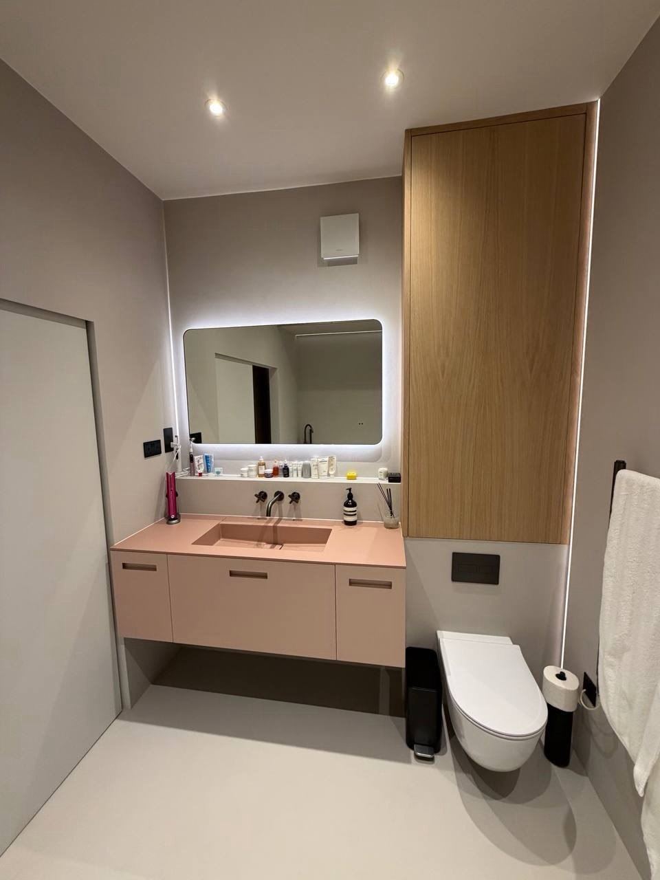 A microcement bathroom with a pink integrated basin and vanity, backlit mirror and a full-height oak cabinet