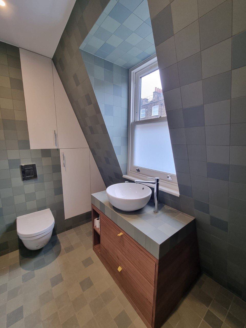 A dormer bathroom tiled floor to ceiling in mixed green square tiles, with a tiled-top walnut vanity
