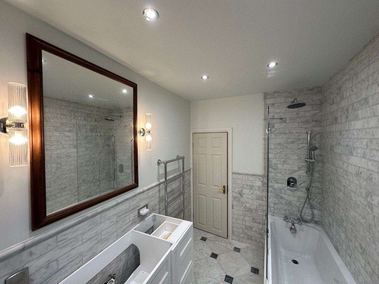 A bathroom in Carrara marble brick tiles with a marble floor, a bath with a glass screen and a large timber mirror